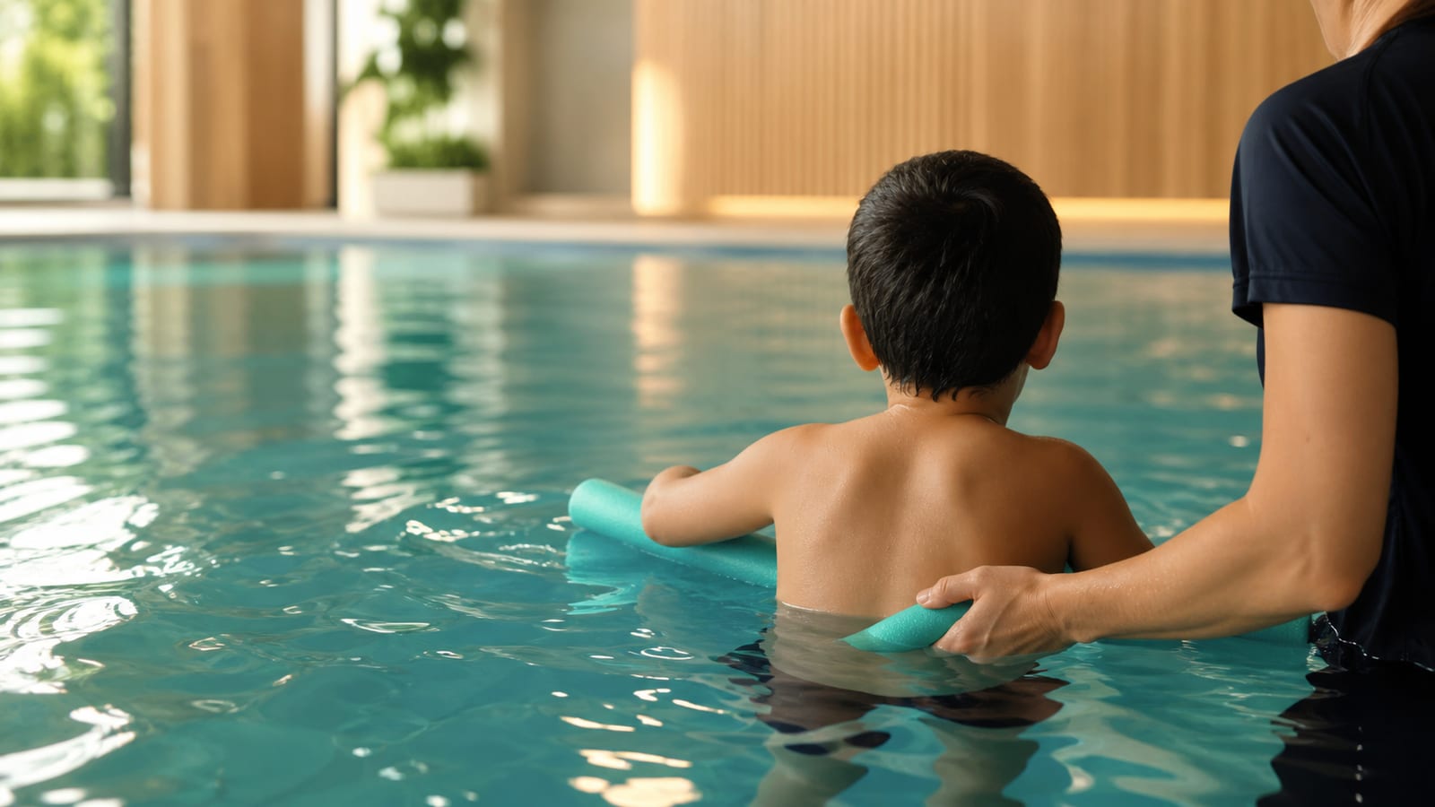 Child in a therapeutic swimming lesson with a supportive instructor in a pool, photographed from behind with no identifiable face.